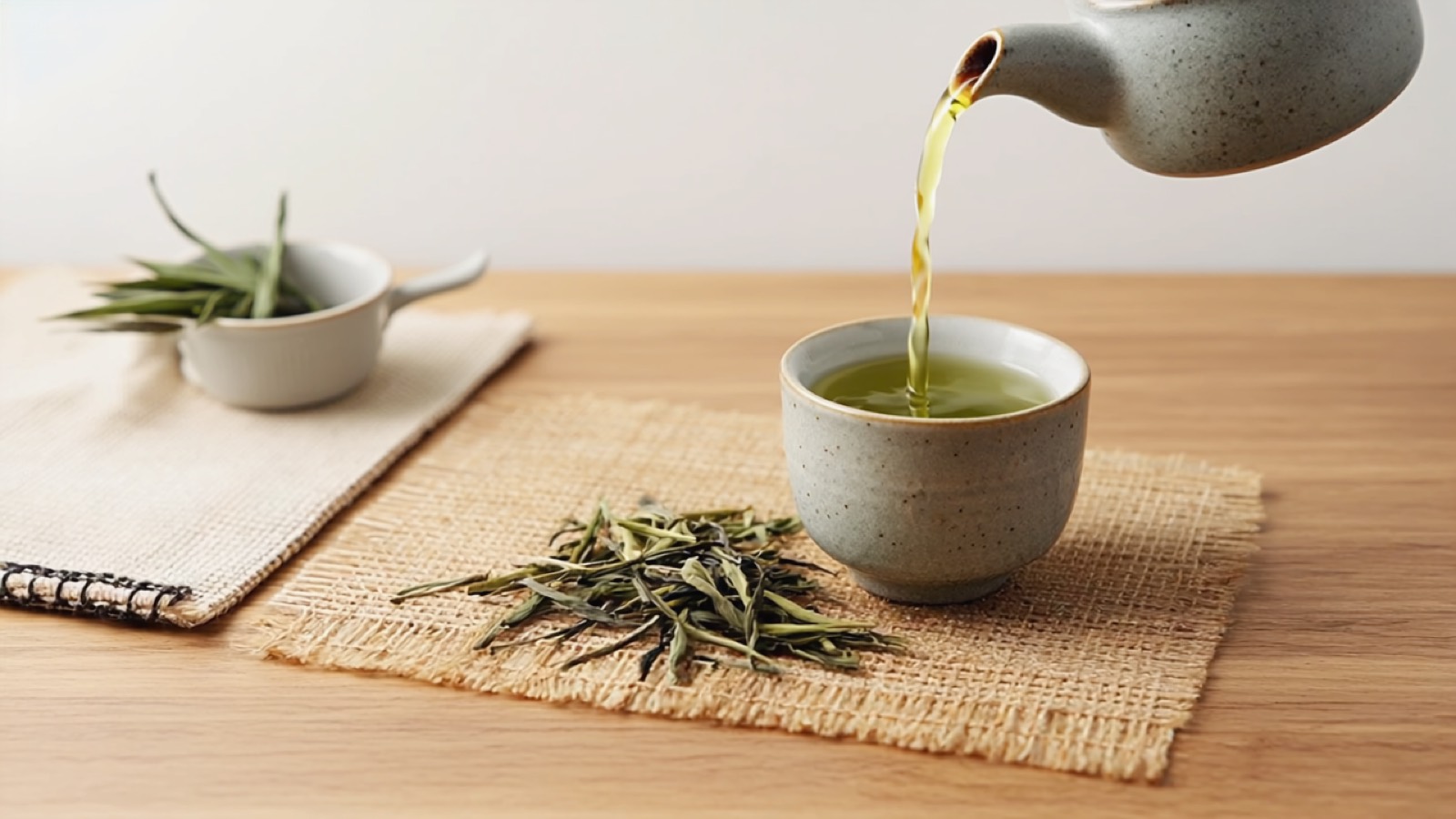 Eine kleine japanische Kyusu-Teekanne gießt hellgrünen Sencha in eine henkellose Schale, daneben lose grüne Teeblätter auf einem warmen Holztisch in weichem Tageslicht.
