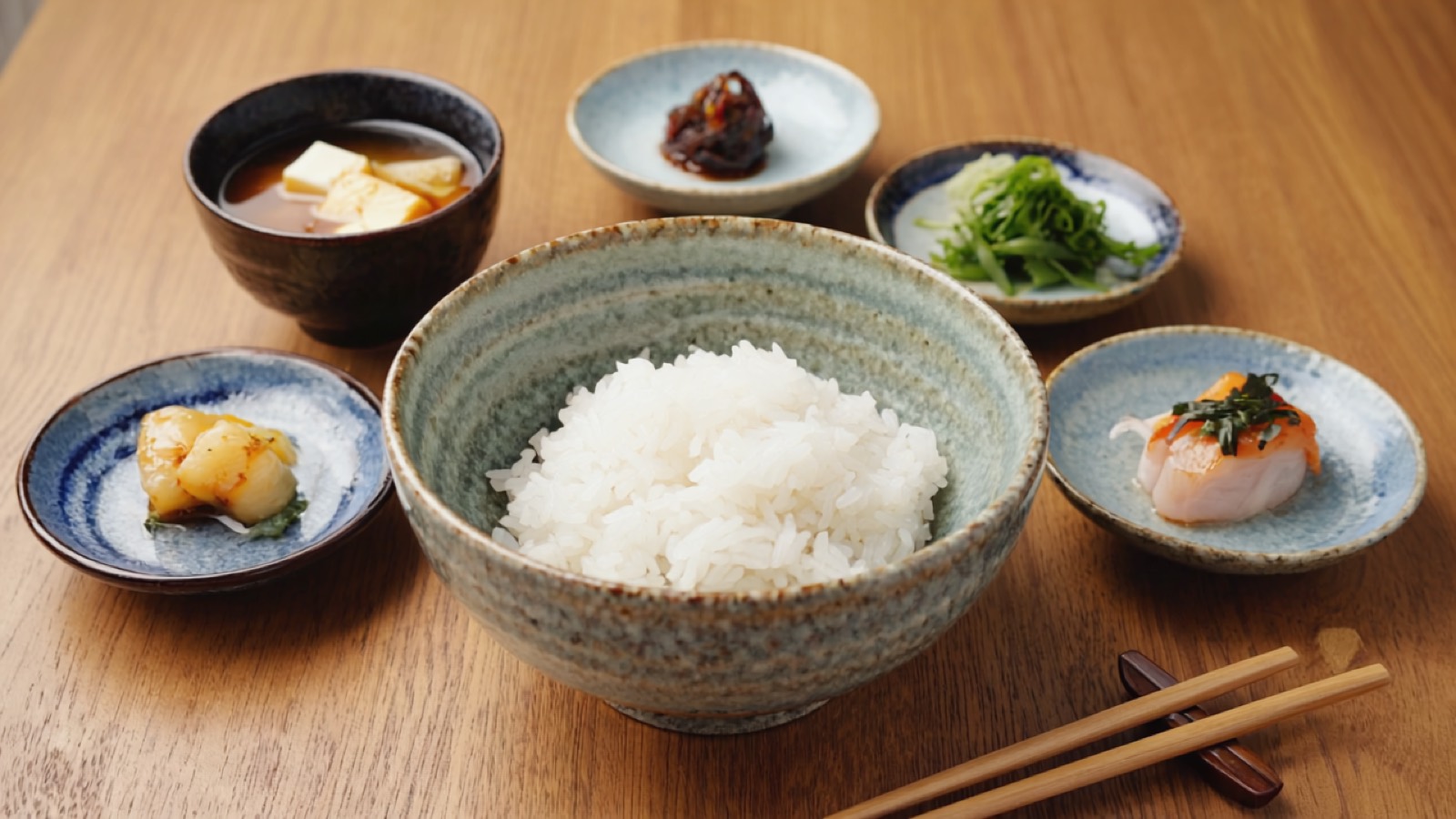 Ein japanisches Alltagsgedeck von oben — eine Schale weißer Reis in der Mitte, Misosuppe und mehrere kleine Beilagen auf handgemachtem Keramikgeschirr in warmem Tageslicht.